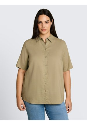 Femme aux longs cheveux foncés portant une chemise beige ample à manches courtes boutonnée et un jean bleu sur un fond uni.