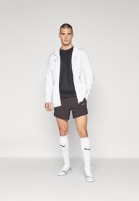 Hvid zip-up hoodie med røde accenter, sort t-shirt, sorte shorts og hvide knæhøje sokker. Modellen har sportssko på med farverigt design.