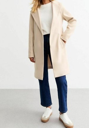 Femme portant un manteau beige longueur genou, un pull blanc, un jean droit bleu marine et des baskets blanches, debout avec une main dans la poche.