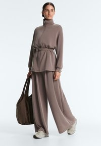 Nainen pukeutuneena taupe-värisiin levenevälahkeisiin housuihin ja vastaavaan soljella vyötettyyn vetoketjutoppiin, pitäen suurta ruskeaa olkalaukkua, seisten yksivärisen taustan edessä.