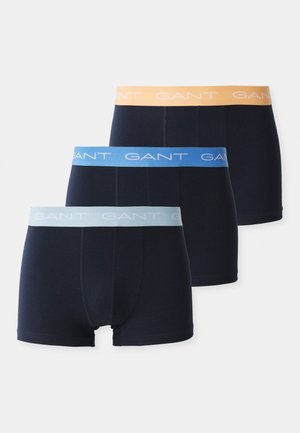 Trois paires de boxer briefs pour hommes en bleu marine, avec des ceintures élastiques bleu clair, bleu foncé et orange sur lesquelles le nom de la marque "GANT" est imprimé.