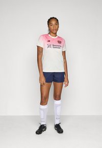 Soccer jersey in light cream with pink gradient, featuring logos. Navy shorts, white knee socks with padding, and black cleats.