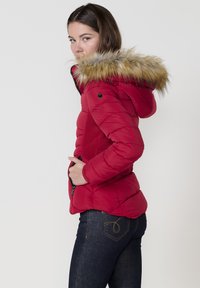 Raudonas pūkinis švarkas su dirbtinio kailio gaubtu, siuvinėta konstrukcija, šoninės kišenės ir juodas užtrauktukas. Dėvimas su tamsiai mėlynomis džinsomis, turinčiomis dekoratyvinę siuvinėjimą.