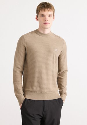 Brown knitted sweater with a high neck, ribbed cuffs, and hem. Features a small logo patch on the chest. Paired with black trousers.