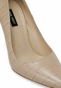 Scarpa con tacco alto beige in pelle con motivo cocodrillo testurizzato, caratterizzata da una punta appuntita e una fodera interna liscia. Etichetta del brand visibile all'interno.