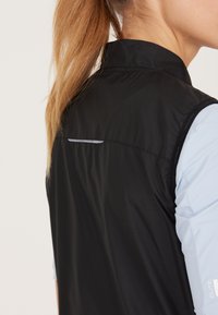 Schwarze Weste mit hohem Kragen, ausgestattet mit einem reflektierenden Akzent auf der Rückseite und hellblauen Ärmel-Einsätzen. Hergestellt aus glattem, leichtem Material.
