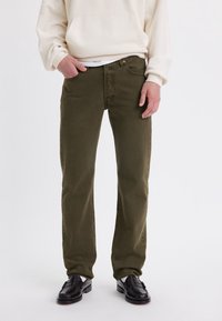 Pantalones de algodón color verde oliva con corte de pierna recta, diseño clásico de cinco bolsillos y cierre de botón en la cintura. Combinados con zapatos negros.