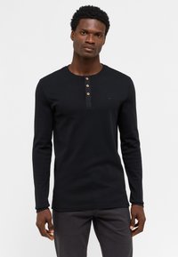 Camisa henley de manga larga negra con cuello redondo, tres botones de madera y un pequeño logo en el lado izquierdo del pecho, confeccionada en tela de canalé.