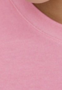 Vaaleanpunainen kangas, jossa on pehmeä tekstuuri, pyöreä pääntie ja näkyvät ompelusaumat. Lähikuva korostaa väriä ja materiaalia.