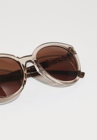Ochelari de soare maro cu un cadru din plastic transparent, lentile rotunde largi și un model asemănător unei lanțuri pe tije.