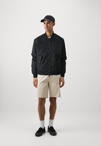 Svart bomberjakke med glidelås, to frontlommer, brukt over en hvit skjorte, kombinert med beige shorts og svarte slip-on sko.