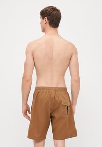 Brune svømmebukser laget av et glatt, lett materiale; har en elastisk midjebånd og en sidelomme med glidelåsdetalj.