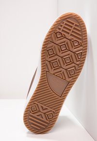 Hnědá gumová podrážka s texturovaným vzorem a hexagonálními tvary. Bílá mezipodešev s hladkým povrchem. Logo Converse viditelné na patě.