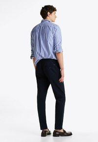 Camicia a righe blu e bianche con maniche arrotolate, abbinata a pantaloni blu scuro e mocassini marroni, vista da dietro.