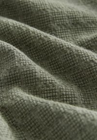 Tkana tkanina w stonowanym zielonym kolorze, z teksturowanym wzorem z przeplatającymi się kwadratami. Miękka, nieco szorstka w dotyku, o bogatej wizualnej głębi.