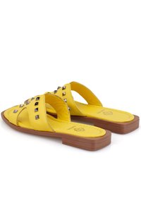 Gelbe flache Sandalen mit gekreuzten Riemen, die mit silbernen Nieten verziert sind. Hergestellt aus synthetischem Material mit einer flachen braunen Sohle und gepolstertem Fußbett.