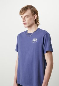 Camiseta de algodón morada con mangas cortas, que presenta un pequeño logo blanco en el pecho. Textura suave y corte relajado.