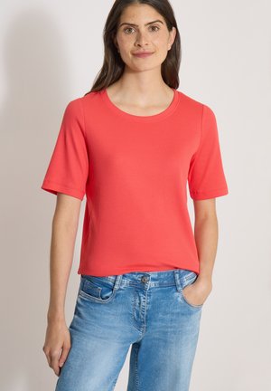 Ung kvinde iført en simpel rød kortærmet trøje og lyseblå jeans, stående med den ene hånd i lommen mod en ensfarvet baggrund.