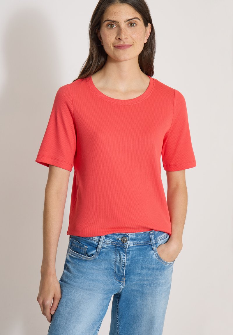 Jeune femme portant un t-shirt rouge uni à manches courtes et un jean bleu clair, debout avec une main dans la poche sur un fond uni.