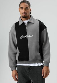 Grijs-zwarte sweatvest met ritssluiting en kraag, met een wit logo op de borst en geribbelde manchetten en zoom.