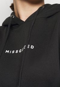 Černá mikina s kapucí vyrobená z měkké tkaniny, se přední kapsou a bílým nápisem "MISSGUIDED" přes hruď.