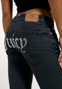 Κοντινό πλάνο ατόμου που φοράει μαύρο τζιν Juicy Couture με γράμματα από στρας "Juicy" και δερμάτινη ετικέτα με το λογότυπο στη μέση.