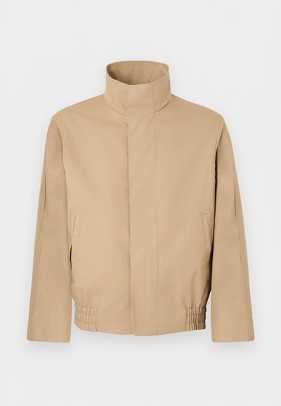 Veste beige à col montant avec manches longues, fermeture avant cachée, poches latérales et taille élastiquée sur fond blanc uni.