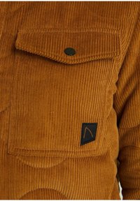 Poche de veste en velours côtelé marron avec rabat sécurisé par un bouton noir et une petite étiquette en tissu noire avec un motif orange cousu.