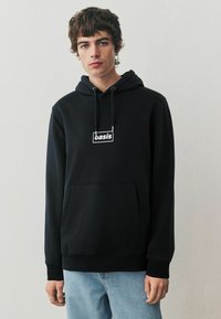 Zwarte hoodie van katoen met een voorzak, voorzien van een wit rechthoekig logo. Het ontwerp heeft een capuchon met trekkoord en ribgebreide manchetten.