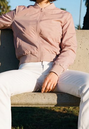 Osoba ubrana w jasnoróżową kurtkę na zamek i białe spodnie, siedząca na betonowej ławce na zewnątrz w słońcu.