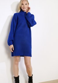 Frau in einem langärmeligen, königsblauen Strickkleid und schwarzen Knöchelstiefeln, die an einer weißen Wand steht.