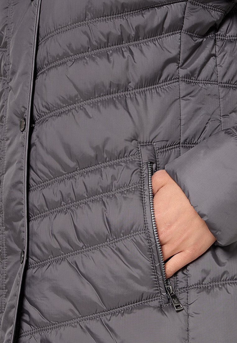 Ręka w kieszeni szarej pikowanej kurtki puchowej z widocznym zamkiem i zatrzaskami z przodu.