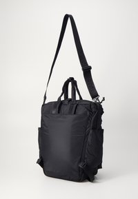 Sac fourre-tout en nylon noir avec bandoulière réglable, poignées supérieures et poches latérales ; présente un design rembourré et une texture lisse.