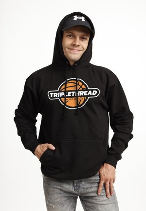 Jonge man die een zwarte hoodie draagt met een basketballogo en de tekst "Triple Thread Hoopwear", een zwarte Under Armour pet en grijze jeans, glimlachend.