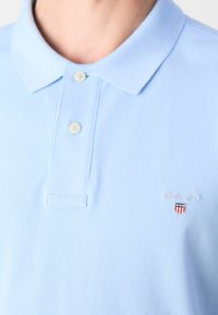 Polo bleu clair avec deux boutons fermés et un petit logo GANT avec emblème en forme de bouclier sur la poitrine.