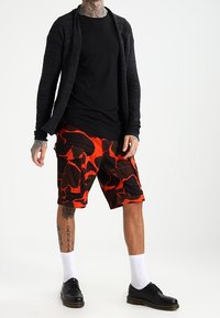 Czarny kardigan z długim rękawem na czarnej koszulce, w połączeniu z pomarańczowo-czarnymi szortami typu cargo w kamuflaż, białymi skarpetkami i czarnymi butami.