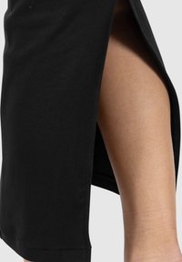 Sort ribstrikket nederdel med højt sidespalte, der afslører bar lår mod en ensartet lys baggrund.