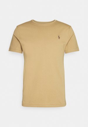 Yksivärinen beige lyhythihainen paita pyöreällä pääntiellä, vasemmassa rinnassa pieni tummanvärinen kirjailtu logo, valkoisella taustalla.