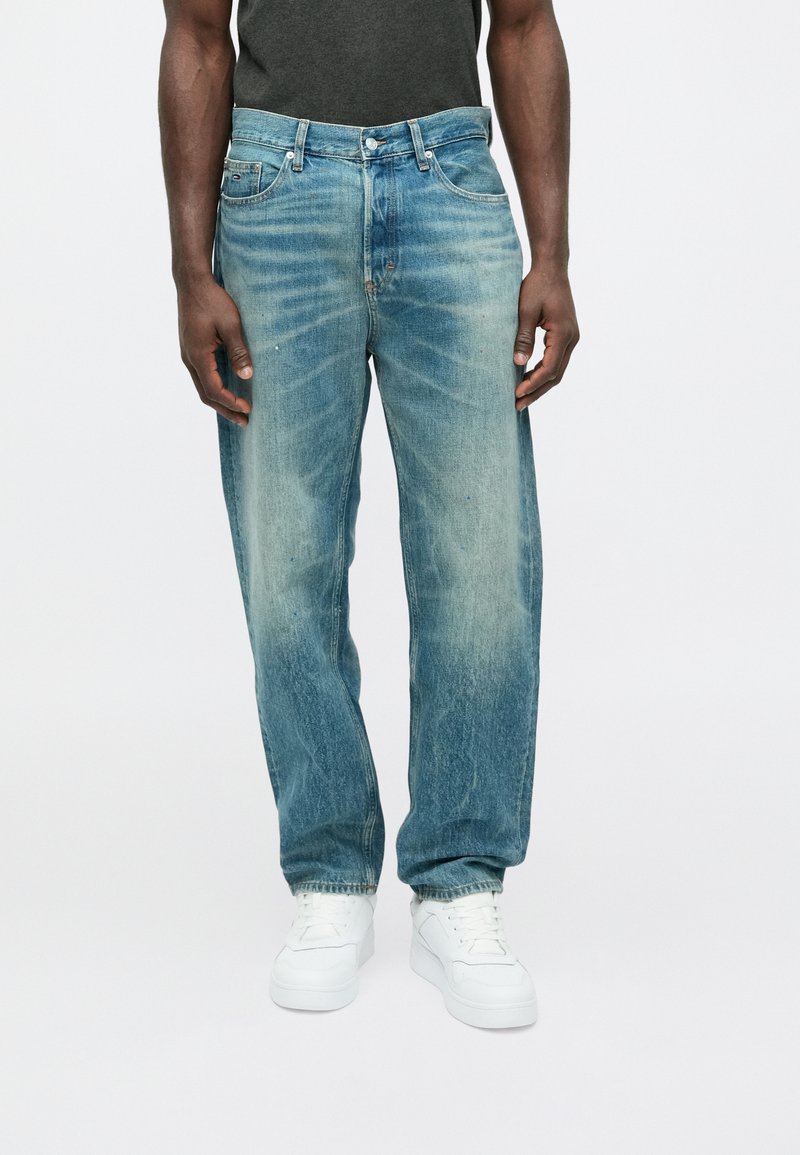 Light blue denim jeans with a faded effect, straight-leg cut, five-pocket design, and metal rivets. Worn with white sneakers.