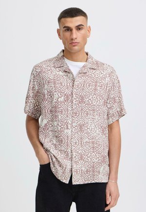 Jeune homme portant une chemise à manches courtes à motifs boutonnée sur un T-shirt blanc, avec une main dans la poche d'un pantalon noir, sur fond uni.