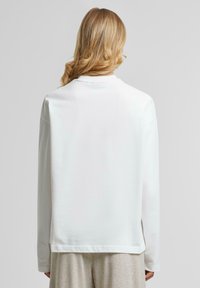 Weißes Langarmshirt aus glattem Material, mit Rundhalsausschnitt und einem Logodetail am Ärmel. Lockere Passform mit geraden Nähten.