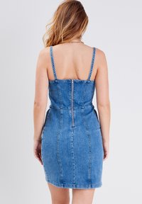 Denimkjole med tynde stropper, tætsiddende liv, og lynlås lukning bagpå. Lyseblå farve med en glat tekstur og sømdetaljer.