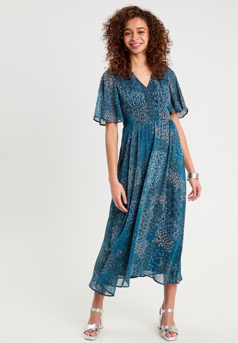 Robe maxi florale teal avec des manches courtes volantées, taille froncée et ourlet fluide. Portée avec des sandales à plateforme argentées.