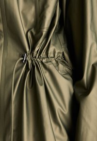 Olive green, water-resistant fabric jacket featuring gathered waist detail with a cinch cord and metallic toggle, smooth texture.