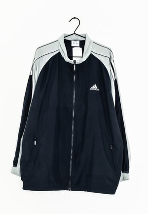Veste de sport Adidas noire et grise avec fermeture éclair intégrale, poches latérales zippées et logo sur la poitrine gauche, suspendue sur un cintre noir.