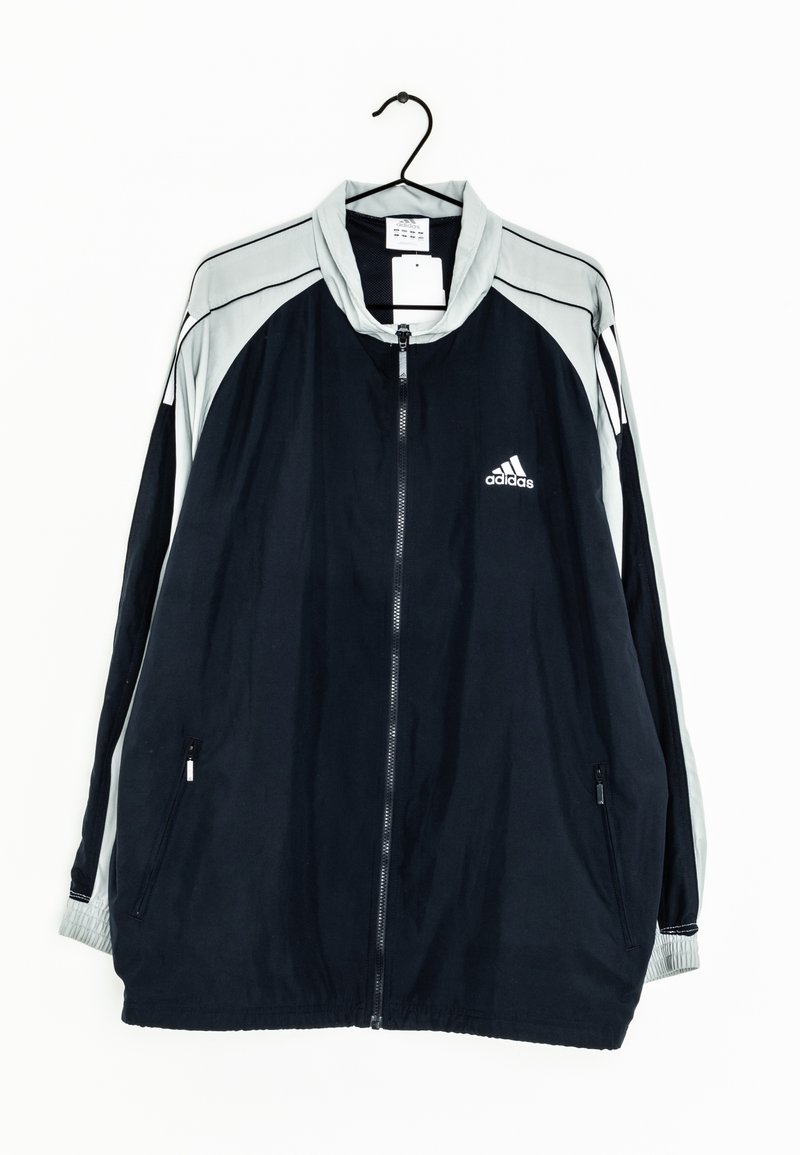 Veste de sport Adidas noire et grise avec fermeture éclair intégrale, poches latérales zippées et logo sur la poitrine gauche, suspendue sur un cintre noir.
