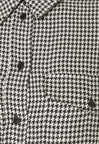 Svartvit houndstooth-mönstrad skjorta med krage, en bröstficka och svarta knappar, tillverkad av strukturerad tyg.