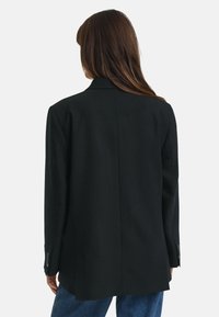 Zwarte, oversized blazer met een gelapt kraag, gestructureerde schouders en een vlakke rugnaad. De stof is textuur met een gladde afwerking.