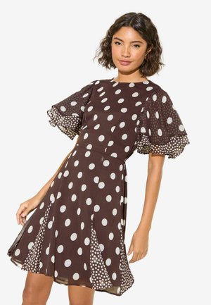 Kobieta ubrana w brązową sukienkę za kolano z dużymi i małymi białymi kropeczkami oraz falbanowymi rękawkami, stojąca na białym tle.
