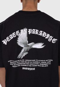 Svart bomull t-skjorte med en hvit grafikk av en due og fet tekst som sier "PEACE IN PARADISE" med inspirerende setninger under.